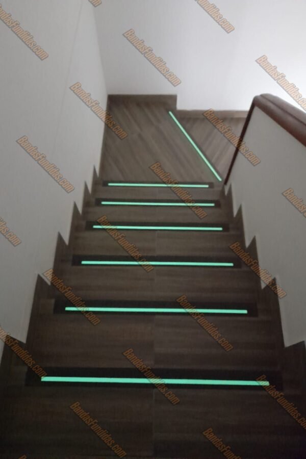 Bandas antideslizantes fundidas con línea fotoluminiscente aplicadas sobre escaleras interiores tipo madera para visibilidad nocturna.